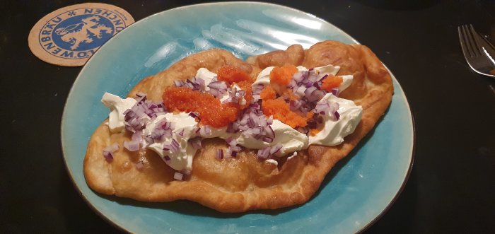 Langos med gräddfil, rödlök och röd kaviar på en blå tallrik.