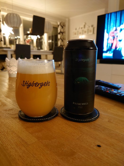 Ett glas öl märkt "Stigbergets" bredvid en ölburk på ett bord, med TV i bakgrunden.