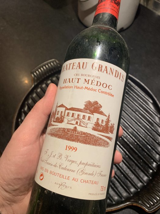 En hand håller en vinflaska märkt "Chateau Grandis Haut Medoc 1999" inomhus.