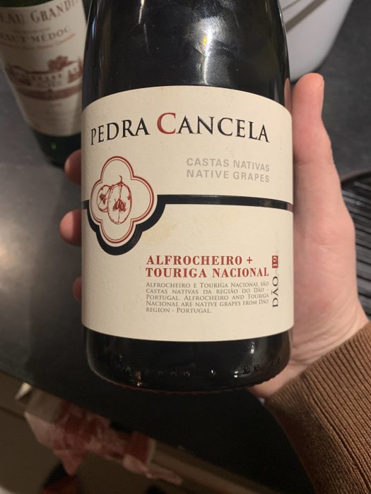 Hand håller en flaska Pedra Cancela vin med etikett som visar druvsorterna Alfrocheiro och Touriga Nacional.