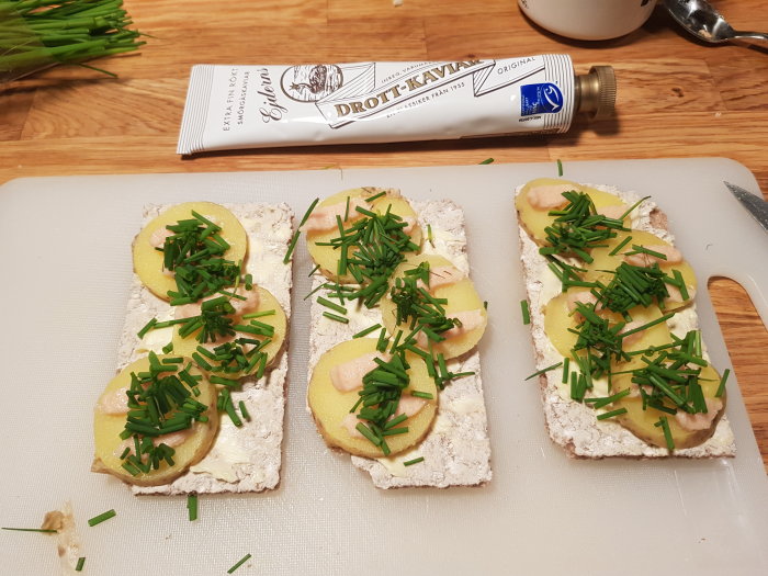 Knäckebröd med smör, skivor av kall kokt potatis, mildrökt kaviar och hackad gräslök på skärbräda.