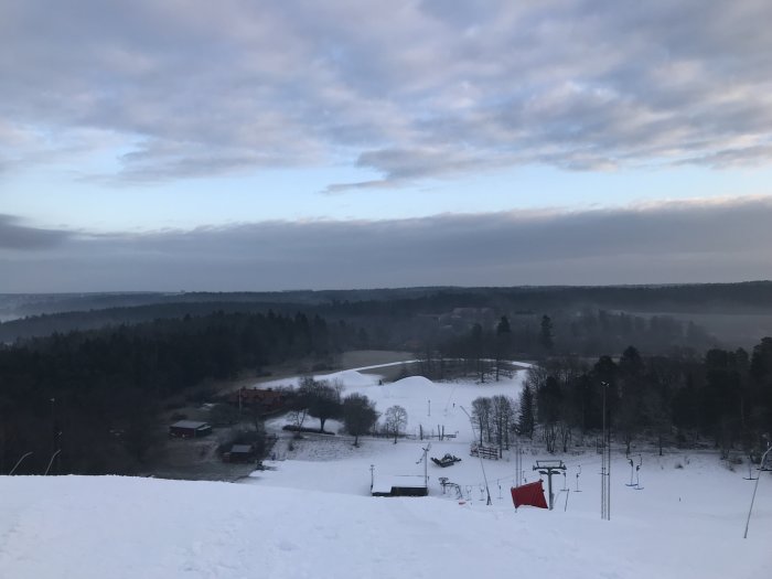 Vy över en snötäckt skidbacke med skidliftar och träd i gryningen.