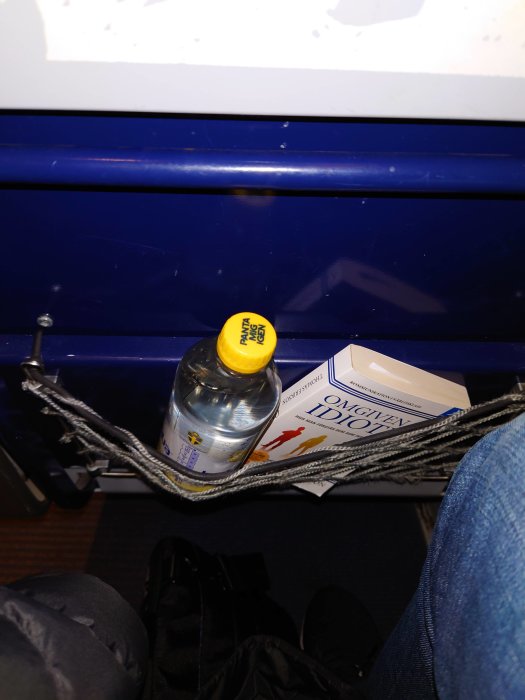 Flaska och bok i ett förvaringsnät på en buss eller tåg, med ett par ben och en väska i förgrunden.
