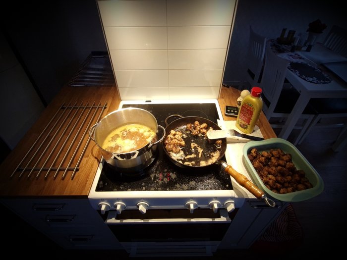 Kök med kastrull och stekpanna på spis, förkokade köttbullar i skål och flaska med senap.