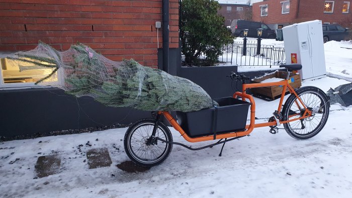 Lastcykel i orangefärg med inslagen julgran i snöigt stadsmiljö.