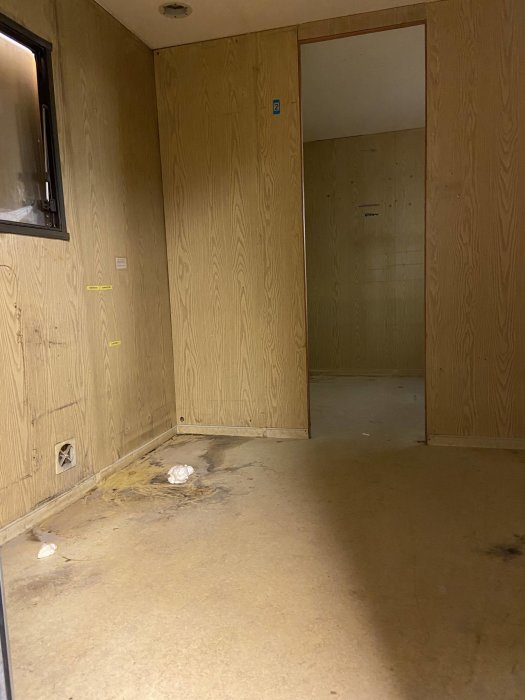 Orenoverat rum med träskivor på väggarna, skadat golv och skräp, i behov av renovering.