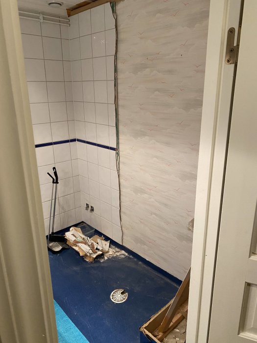 Badrum under renovering med halvt nedrivna väggar och golv, gamla vattenkopplingar synliga i hörnet, rivningsavfall på golvet.