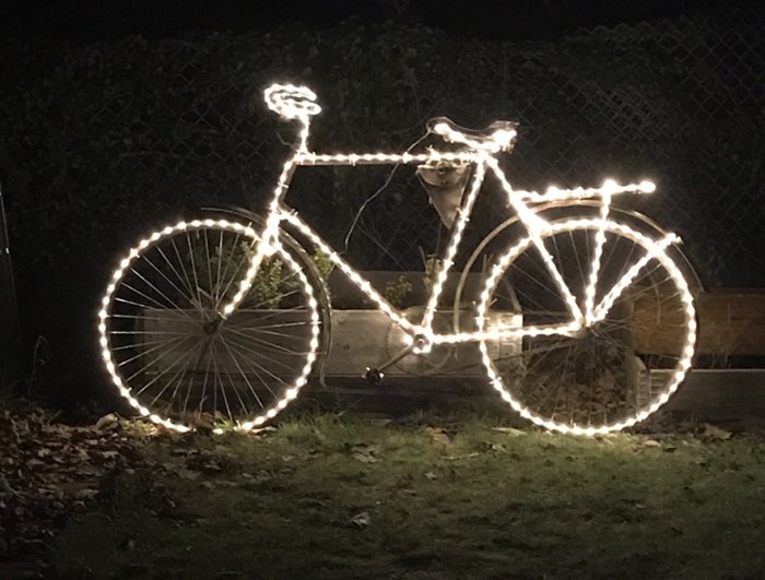 Cykel dekorerad med ljusslingor står på gräs framför en häck på kvällen.