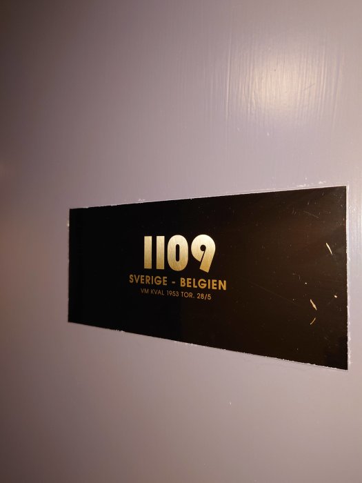 Svart minnesskylt med text "1109 SVERIGE - BELGIEN VM kval 1953 tor. 28/5" på en grå vägg.