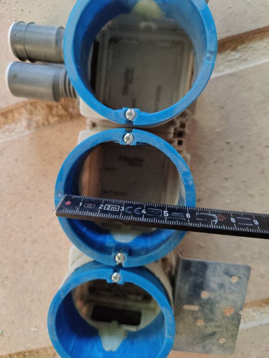Tre blåa elrörsinstallationer i vägg med ett måttband som visar dimensionerna.