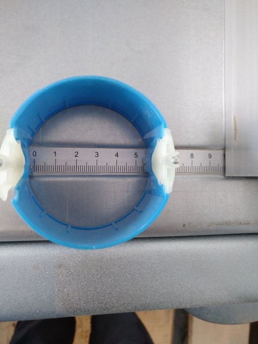 Blå plastklämma med vit märkning ovanpå en metallkant med linjal, visar mätning.