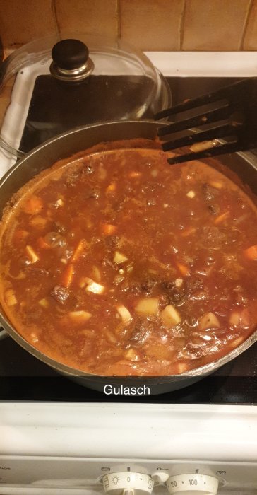 Gulasch på spisen i en svart lergryta med genomskinligt lock, bitar av älg och grönsaker syns.