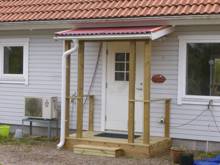 Nybyggd träentré med tak över ytterdörr till vit husfasad och värmepump intill.