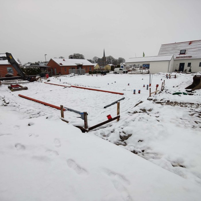 Byggarbetsplats med markarbeten under snötäcke, synliga schakter och märkta rör i förgrunden, med byggnader i bakgrunden.