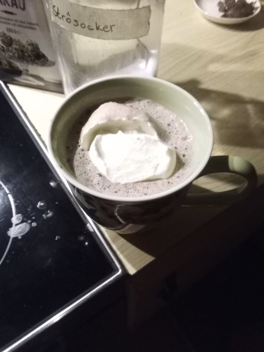 Hemgjord varm choklad med grädde i en kopp, burk med strösocker i bakgrunden.