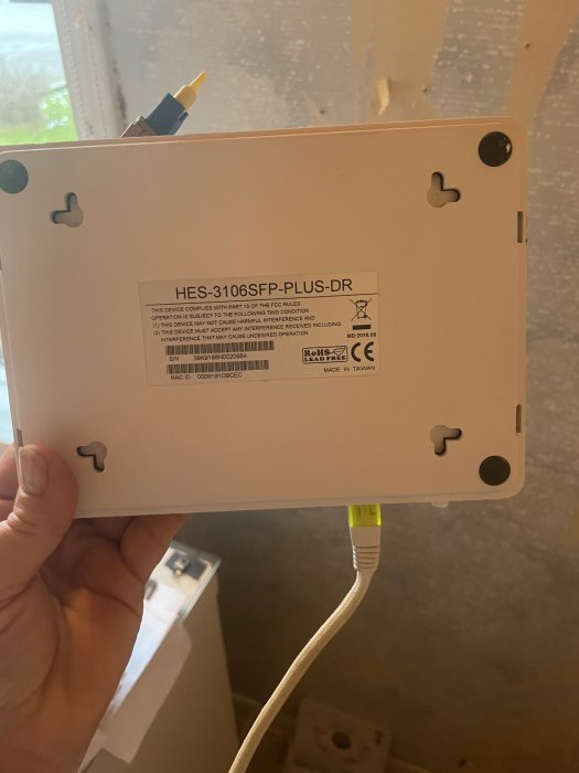 Hand håller en nätverksenhet med modellnummer HES-3106SFP-PLUS-DR och ansluten Ethernet-kabel.