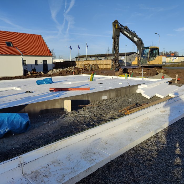 Byggplats med kantelement och cellplast på marken, grävmaskin i bakgrunden under klar himmel.