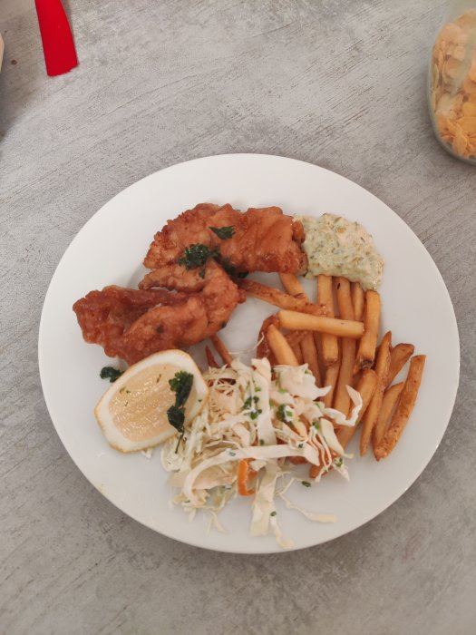Fish and chips på vit tallrik med remouladsås, citronskiva och coleslaw, otydlig bakgrund.