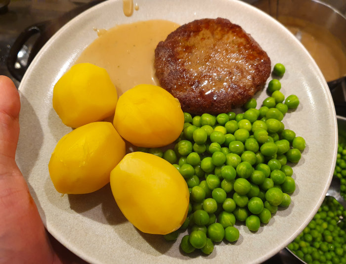 Ett fat med pannbiff gjord på sojafärs, kokta potatisar, gröna ärtor och brunsås hållet i en hand.