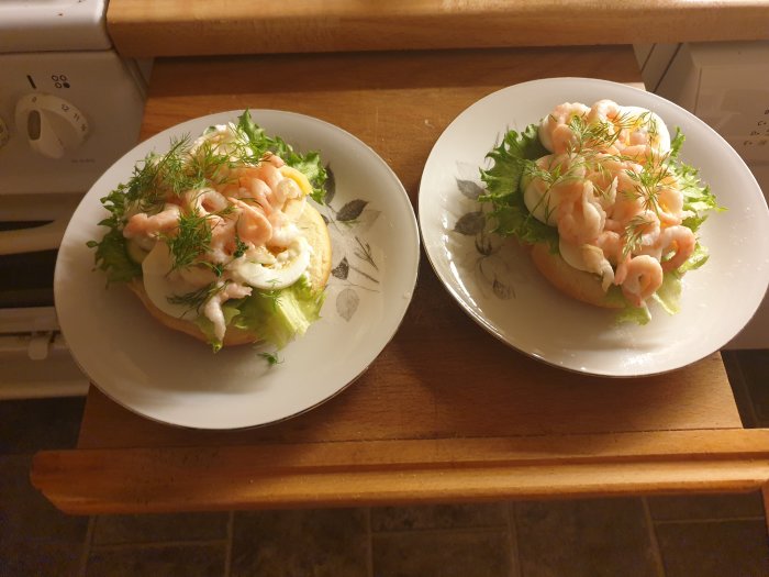 Två räkmackor med sallad och dill på vita tallrikar framför en ugn.