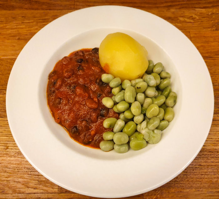 En tallrik med chilligryta, kokt potatis och bondbönor serverad på ett träbord.
