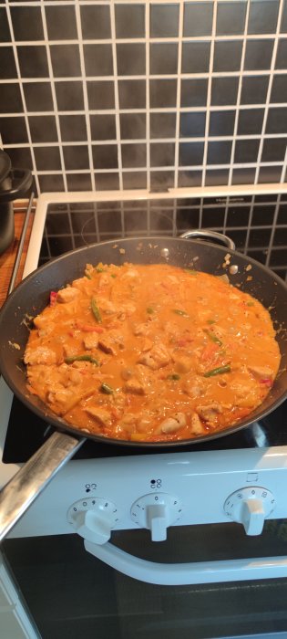 Stekpanna på spis med thailändsk curry och grönsaker som tillagas, kakelvägg i bakgrunden.