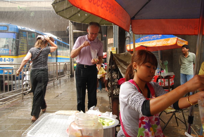 Gatukök i Thailand under regn där en kvinna lagar mat och en man äter under ett paraply.