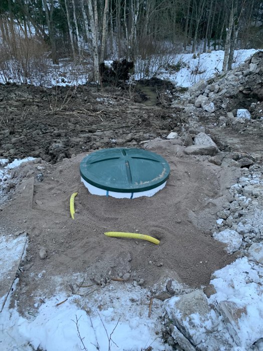 Nyligen nedgrävt avlopp med grön lock och gul slang synlig i utgrävningen med snö och skog i bakgrunden.