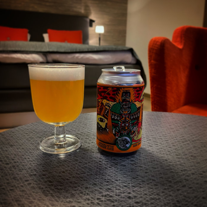 Ölglas med ljus dryck bredvid en färgglad ölburk på ett bord, soffa i bakgrunden.