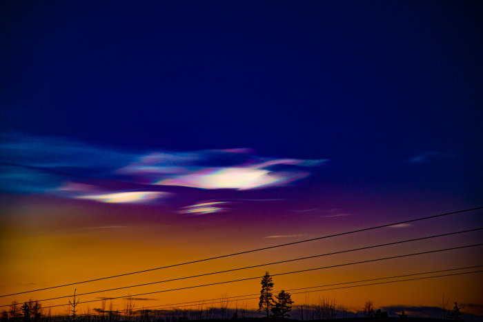 Färgglad himmel vid solnedgång med ovanliga molnformationer och silhuetter av träd och elledningar.