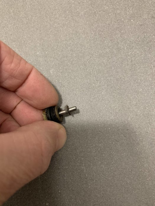 Hand håller en demonterad radiatorventil med synligt stift och ventilkägla, fullt öppen för flöde.