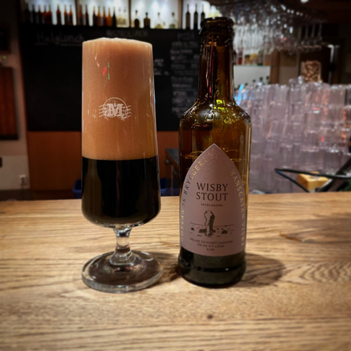 Överfyllt glas med mörkt öl bredvid en tom Wisby Stout-flaska på en bar.