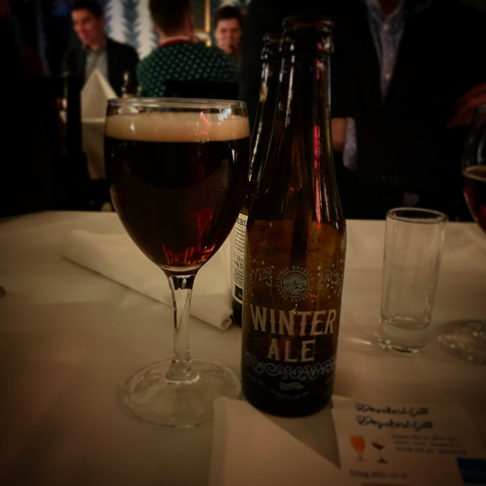 Flaska märkt 'WINTER ALE' och ett ölglas på ett bord med suddig bakgrund av människor.