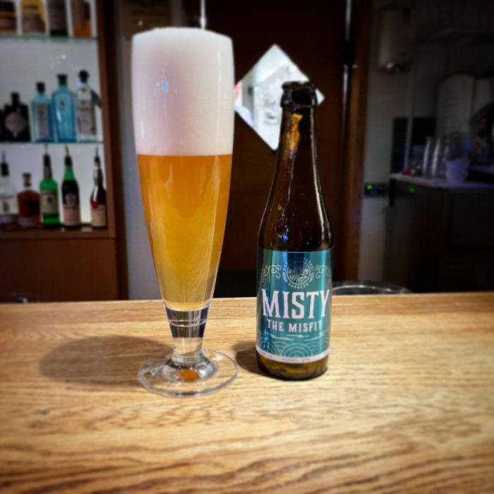 Ölglas med överflödigt skum bredvid en ölflaska på en träyta, suddig bakgrund med flaskor.