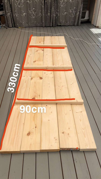 Träplankor utlagda på grått trädäck märkta med mått 330cm x 90cm för byggprojekt.