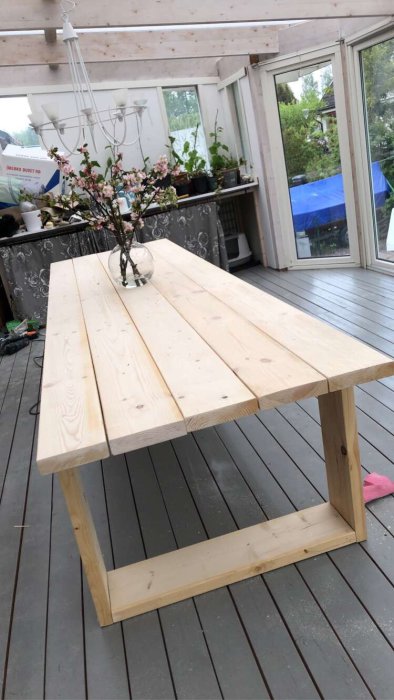 Nybyggt enkelt träbord av plank står på en terrass med en vas blommor ovanpå och inredningsdetaljer i bakgrunden.