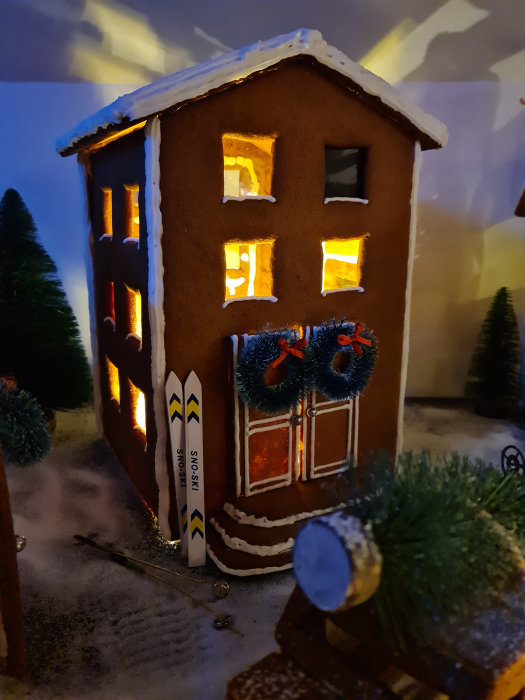 Handgjord pepparkakshusmodell med upplysta fönster och skiddekor vid dörren, omgiven av miniatyrträd och snö.