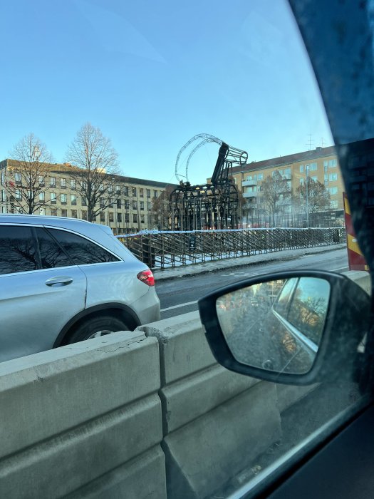 Vy från bilfönster visande en pariserhjulskonstruktion i en stadsmiljö omgiven av byggnader.