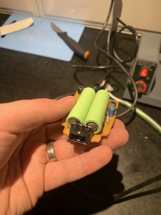 Hand holding an electronic circuit board with two new green batteries, knife, and charger in the background.