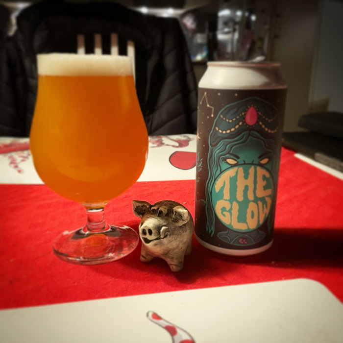 Ett glas öl bredvid en ölburk med texten "THE GLOW" och en liten grishälsningsfigur framför på en röd duk.