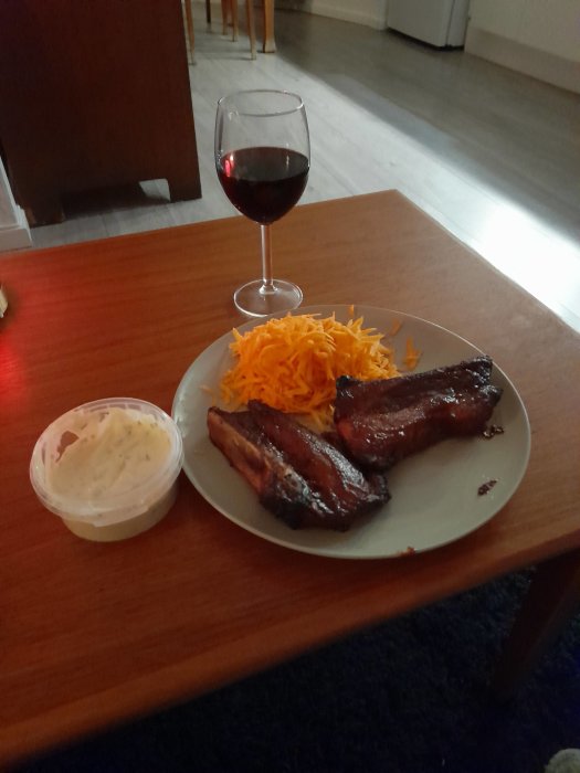 Grillade klubbor på tallrik med rivna morötter, köpesås och ett glas rött vin på ett vardagsrumsbord.