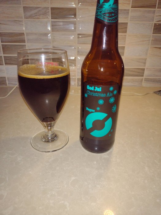 Öppnad julölflaska bredvid ett glas fyllt med mörk öl på ett köksbord.