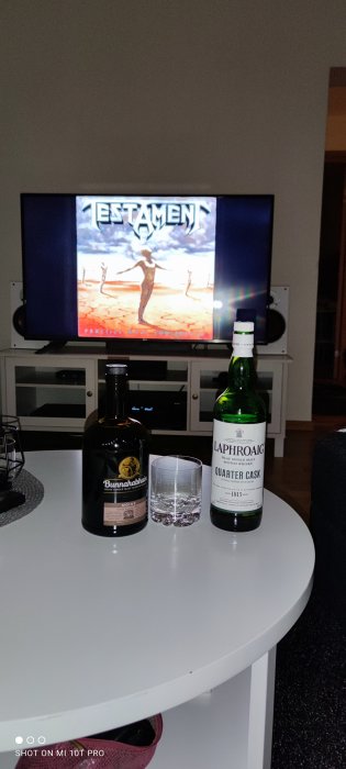 TV-skärm som visar albumomslaget för "Testament", med två whiskflaskor och ett glas på bordet framför.