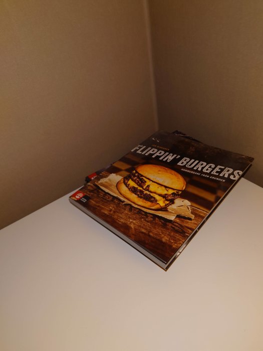 En bok med titeln "Flippin' Burgers" ligger på ett vitt bord med en bild av en hamburgare på omslaget.