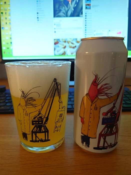 Ölglas med konstnärligt tryck framför en matchande ölburk, placerade på ett skrivbord med datorskärm i bakgrunden.