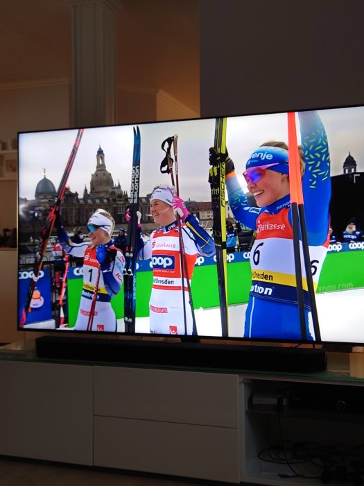 Tre skidåkare firar på prispallen visade på en TV-skärm, med arkitektur i bakgrunden.