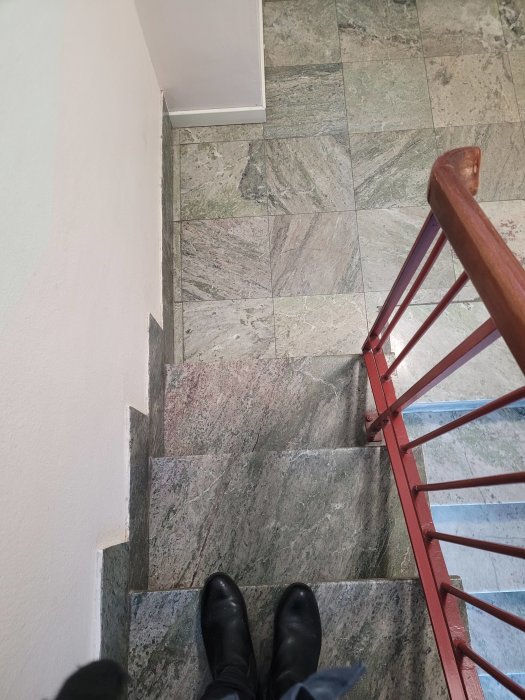 Person står överst på en trappa med marmorerade klinkerplattor och röda metallräcken.