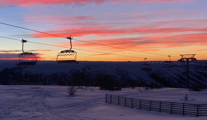 Skidliftar mot en dramatisk solnedgångshimmel över ett snötäckt landskap.