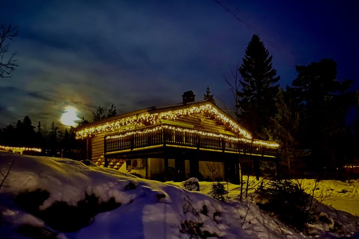 En timmerstuga med upplyst veranda i en snöklädd omgivning under kvällstid.