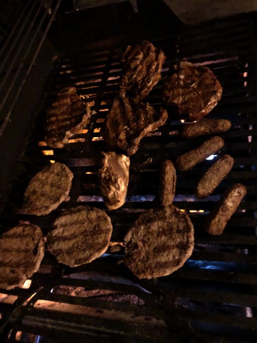 Stekande kött och korvar på en mörk grill med synliga lågor under.